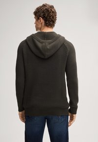 Donkergroen, gebreide trui met capuchon en een textuurpatroon, met geribde manchetten en zoom. Gekleed met blauwe denim jeans, gezien vanaf de achterkant.