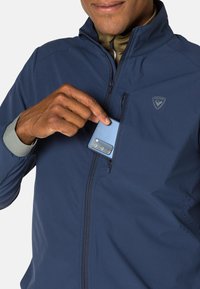Dunkelblaue Jacke mit einer Reißverschlusstasche auf der Brust, die ein hellblaues Smartphone hält; aus glattem, wasserabweisendem Material gefertigt.