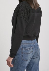 Femme portant un haut noir à manches longues avec des épaules en dentelle et un jean en denim bleu, vue de dos sur un fond clair uni.