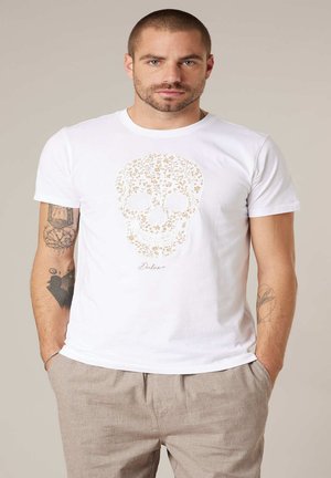 Mann mit kurzen Haaren und Tätowierungen trägt ein weißes T-Shirt mit floralem Totenkopf-Design und beige Hosen, steht mit den Händen in den Taschen.