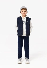 Gilet imbottito blu navy sopra una felpa bianca con logo di coccodrillo, abbinato a pantaloni blu navy e sneakers grigie. Il ragazzo indossa un berretto grigio.