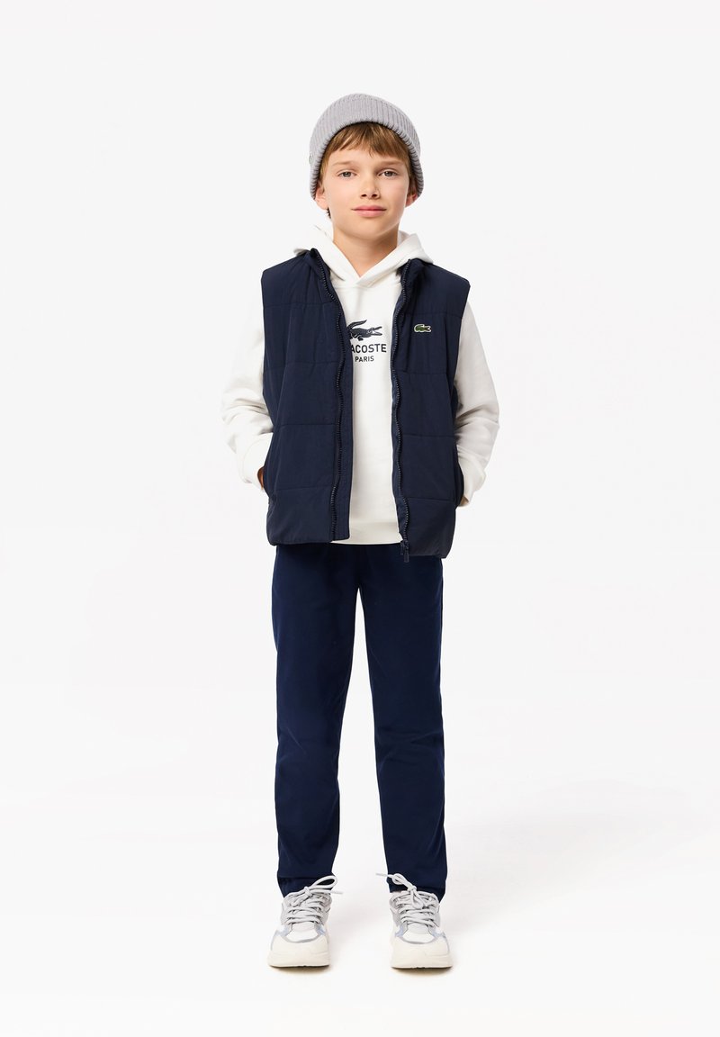 Gilet imbottito blu navy sopra una felpa bianca con logo di coccodrillo, abbinato a pantaloni blu navy e sneakers grigie. Il ragazzo indossa un berretto grigio.