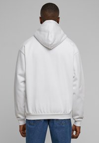 Hvit hettegensere med avslappet passform, subtil tekstur og ribbede mansjetter og kant, paret med blå jeans, sett fra bak.