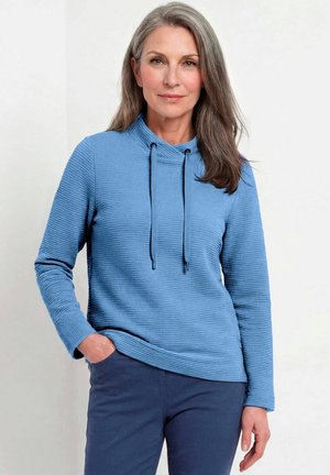 Vrouw met lang grijs haar draagt een blauw getextureerd longsleeve top met koordjes en donkerblauwe broek, staat met één hand in de zak.
