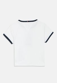 Top alb cu mâneci scurte, cu margine albastru închis. Textură nervurată, design cu decolteu în V, prevăzut cu o etichetă mică cu dungi roșii și albastre în spate.