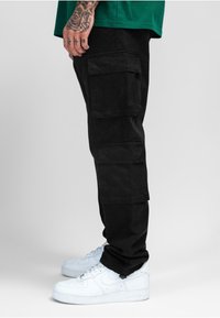Pantalon cargo noir en tissu, doté de plusieurs grandes poches, coupe ample, porté avec des baskets blanches. Détail de texture minimal visible.