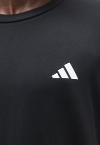 Μαύρο πουκάμισο με λευκό λογότυπο Adidas στο αριστερό στήθος, που δείχνει το γιακά και το πάνω μέρος του στήθους ενός ατόμου.