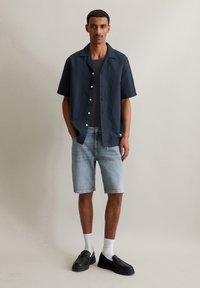 Mariene korteknelleshirt met knopen, gedragen over een zwart T-shirt, gecombineerd met lichtblauwe denim short en zwarte instapschoenen.