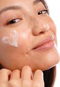 Crema idratante applicata a forma di cuore sulla guancia di una donna, che mostra una pelle liscia con lentiggini e un'espressione rilassata.