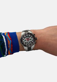 Stainless steel watch on a wrist, featuring a black dial, red accents, Arabic numerals, and a patterned fabric sweater sleeve.