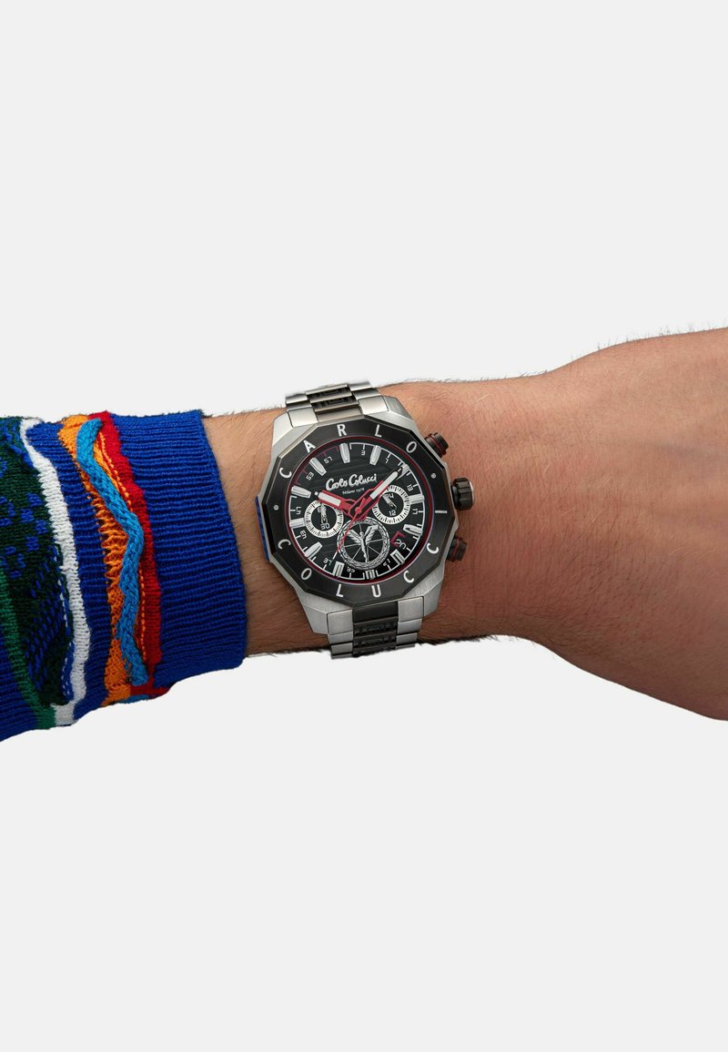 Stainless steel watch on a wrist, featuring a black dial, red accents, Arabic numerals, and a patterned fabric sweater sleeve.