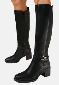 Bottes en cuir noir jusqu'au genou avec un talon carré, fermeture éclair et accent de boucle à la cheville. Texture lisse, silhouette structurée.