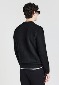 Mann mit lockigem dunklem Haar, der schwarze Sonnenbrillen, einen schwarzen Pullover über einem weißen Hemd und schwarze Hosen trägt, von hinten auf einem weißen Hintergrund gesehen.