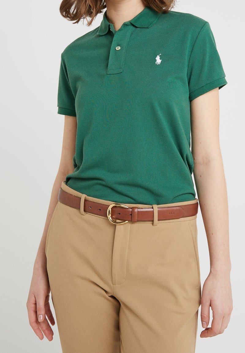 Grön polo med två knappar och en vit logotyp på bröstet, tillsammans med beige byxor och ett brunt bälte med en guldsfärgad spänne.