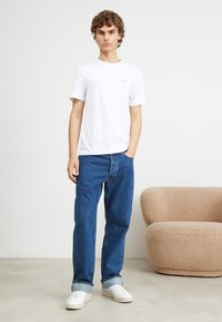 White cotton t-shirt with short sleeves, paired with blue denim jeans and white sneakers. Neutral background with a textured beige sofa.