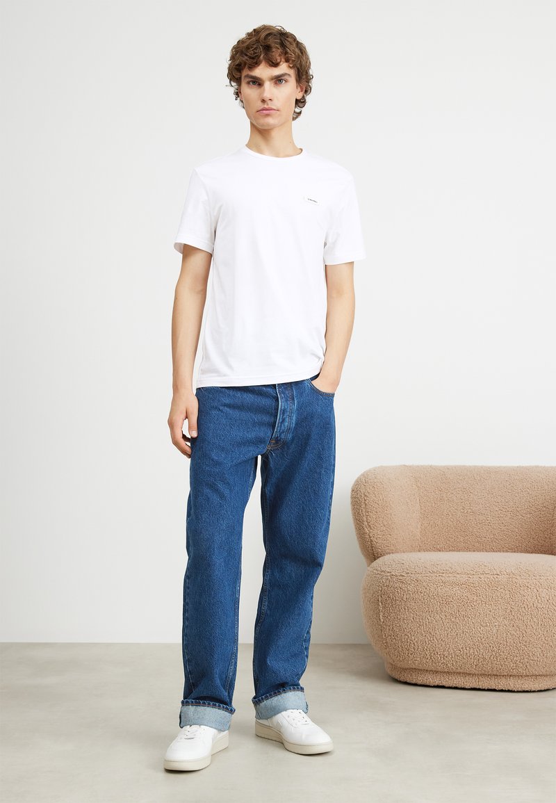 Weißes Baumwoll-T-Shirt mit kurzen Ärmeln, kombiniert mit blauen Denim-Jeans und weißen Sneakern. Neutraler Hintergrund mit einem strukturierten beigefarbenen Sofa.