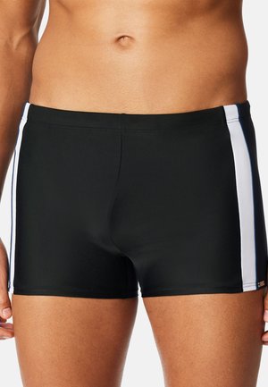 Męski tors ubrany w czarne spodenki kąpielowe z białymi pionowymi paskami po bokach, stojący na jasnym, jednolitym tle.