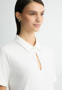Bluză albă cu guler rotund, accentuată cu mărgele de-a lungul gulerului și cu o deschidere în formă de cheie în față. Material neted, mâneci scurte.