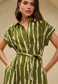 Abito a camicia a righe verdi e crema con maniche corte, chiusura frontale con bottoni e cintura in vita. Il tessuto presenta una finitura texturizzata.