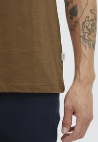 Brun bomull T-skjorte med en buet hem, med et lite merkelapp på siden, kombinert med mørkeblå bukser og en tatovert arm.