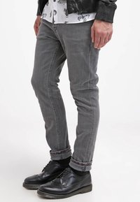 Jeans de mezclilla gris con un corte ajustado, que presentan puños enrollados y un ligero desgaste, combinados con zapatos negros de cordones y una camisa de patrón.