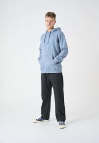 Felpa con cappuccio azzurro chiaro, dotata di tasca a marsupio, cordoncino e piccolo logo decorativo. Abbinata a pantaloni neri e sneakers bianche.