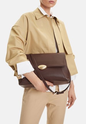 Person in beige jacket holding a large dark brown leather handbag with gold clasp and shoulder strap against white background.