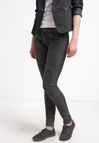Schwarze Denim Skinny Jeans mit glatter Textur, ausgestattet mit Knie-Patches, schlanker Passform und mittlerer Leibhöhe. Kombiniert mit lässigen schwarzen Sneakers.