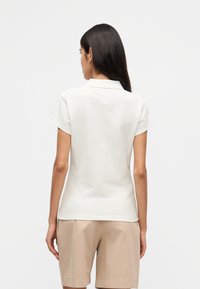 Λευκό πόλο πουκάμισο με κοντά μανίκια, κλασικό γιακά και εφαρμοστή σχεδίαση, συνδυασμένο με μπεζ σορτς. Λεπτή υφή υφάσματος, μινιμαλιστικό λογότυπο.