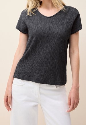 Femme portant un haut noir texturé à manches courtes et un pantalon blanc, visible des épaules aux hanches sur un fond uni.