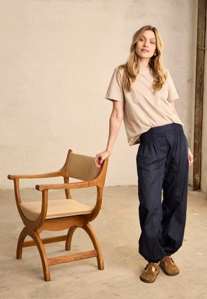 Femme aux longs cheveux blonds appuyée sur une chaise en bois avec un coussin beige, portant une chemise beige, un pantalon noir et des chaussures marron sans lacets.