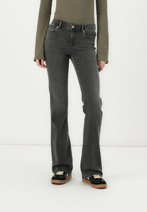 Osoba ubrana w ciemnoszare dżinsy bootcut, czarne trampki z brązowymi podeszwami oraz oliwkowozielony top z długim rękawem, stojąca na białym tle.