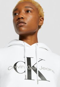 Felpa bianca con cappuccio caratterizzata da un prominente logo nero "CK" e il testo "Calvin Klein Jeans", con hardware dei lacci argentato.