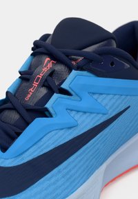 Zilā un tumši zila sporta kurpe ar teksturētu virsmu, gludām gumijas detaļām, kontrastējošu swoosh dizainu un redzamām šnurēm.