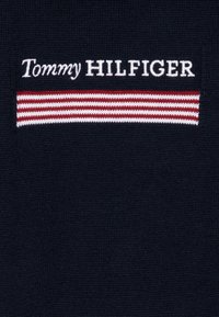 Jūras spēku adījuma audums ar "Tommy HILFIGER" logo baltā krāsā, kuru papildina sarkana un balta svītra virs teksta.
