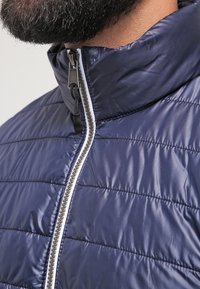 Giacca imbottita blu navy con colletto alto, caratterizzata da linee di trapuntatura orizzontali, una zip frontale con denti argentati e dettagli bianchi a contrasto.