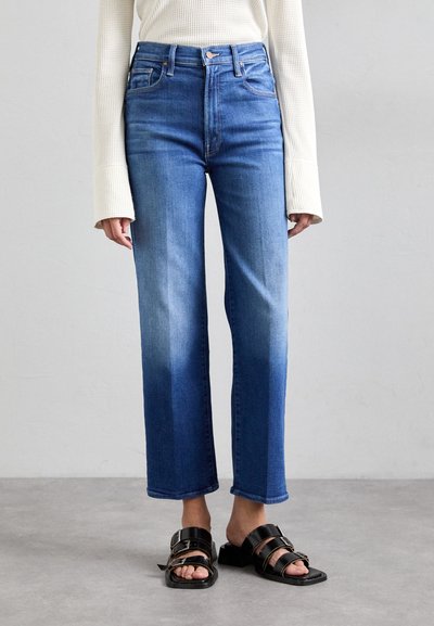 Jean bleu taille haute, jambes droites, avec un dégradé allant du denim foncé au denim clair. Porté avec des sandales noires à semelle plate et un haut blanc côtelé.