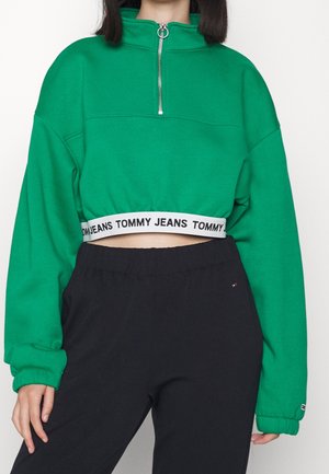 Frau trägt einen grünen, kurz geschnittenen Pullover mit Frontreißverschluss und weißem elastischem Band mit der Aufschrift "Tommy Jeans", kombiniert mit schwarzen elastischen Bundhosen.