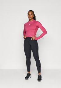Rózsaszín, hosszú ujjú sportfelső, slim fit szabással, magas gallérral és oldalsó panel bordázatokkal, fekete kompressziós leggings-szel és fekete sportcipőkkel párosítva.