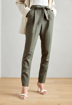 Vrouw draagt olijfgroene high-waist taps toelopende broek met een stoffen riem gestrikt in een strik, witte sandalen met hak en een beige blazer met handen in de zakken.