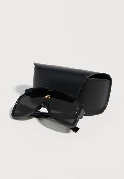 Μαύρα γυαλιά ηλίου με υπερμεγέθη ασπίδα και χρυσή λεπτομέρεια λογότυπου, συνοδευόμενα από μαύρη δερμάτινη θήκη σε λευκή επιφάνεια.