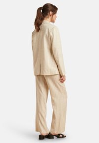 Beige gestreepte pakken set met een relaxed fit. De blazer heeft een enkele split, terwijl de broek een wijde pijp heeft en een zachte textuur. Zwarte blokhak sandalen.