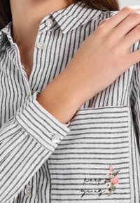Camicia a righe grigia e bianca con tasca decorata con ricami floreali e una sottile texture. Spalline e bottoni perle.