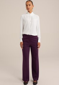 Vit långärmad blus med hög krage, tillsammans med lila vida byxor. Slät tyg och figursydd midjedetalj. Svarta platta skor.