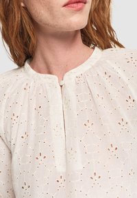 Roodharig persoon die een witte blouse met oogjesborduurwerk en een kleine ronde knoop bij de hals draagt, tegen een effene achtergrond.