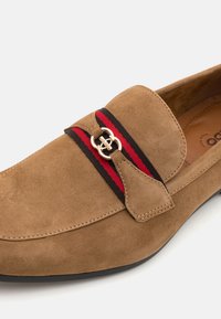 Mocassino in suede color beige con un emblema dorato, fascia decorativa a righe rosse e nere, punta rotonda e suola bassa in gomma nera.