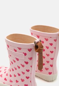 Bottes de pluie en caoutchouc rose avec un motif de cœurs, dotées d'une languette en cuir marron et d'un intérieur texturé, conçues pour le confort et le style.