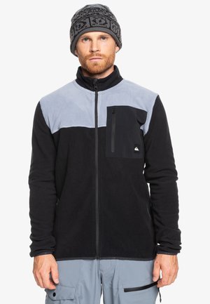 Fleece jacket with black and light gray panels. Features a zippered chest pocket and high collar. Worn with a patterned knit beanie.