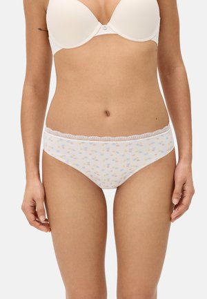 Mujer con sostén blanco y ropa interior blanca con encaje floral, de pie con los brazos relajados contra un fondo liso.