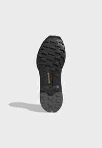 Semelle extérieure en caoutchouc noir avec des rainures hexagonales profondes pour une meilleure traction, avec un logo central orange. Le design est durable et texturé.